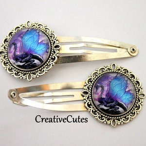 May include: Two silver hair clips with a purple and blue dragon design in a gold filigree frame.  CreativeCutes.