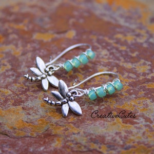May include: Silver dragonfly earrings with light green glass beads. The earrings are hanging from a silver wire.