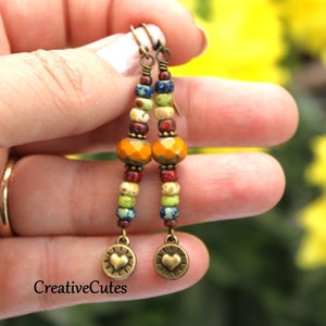 May include: A pair of dangle earrings featuring a heart charm. The earrings are made with colourful beads in shades of red, orange, yellow, green, and blue. The beads are strung on a wire with a bronze-coloured finish.