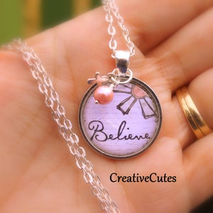 Könnte beinhalten: Eine silberne Kettenkette mit einem runden Anhänger mit einem lila und weißen Blumenmuster und dem Wort "Believe" in schwarzer Schrift. Ein kleiner rosa Perlenanhänger und ein silberner Kreuzanhänger hängen an dem Anhänger.