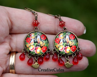 May include: A pair of round, ornate earrings with a floral and bird design. The earrings feature a black background with red, yellow, and white flowers and two birds. Red teardrop-shaped beads dangle from the bottom, and the text "CreativeCutes" is visible.