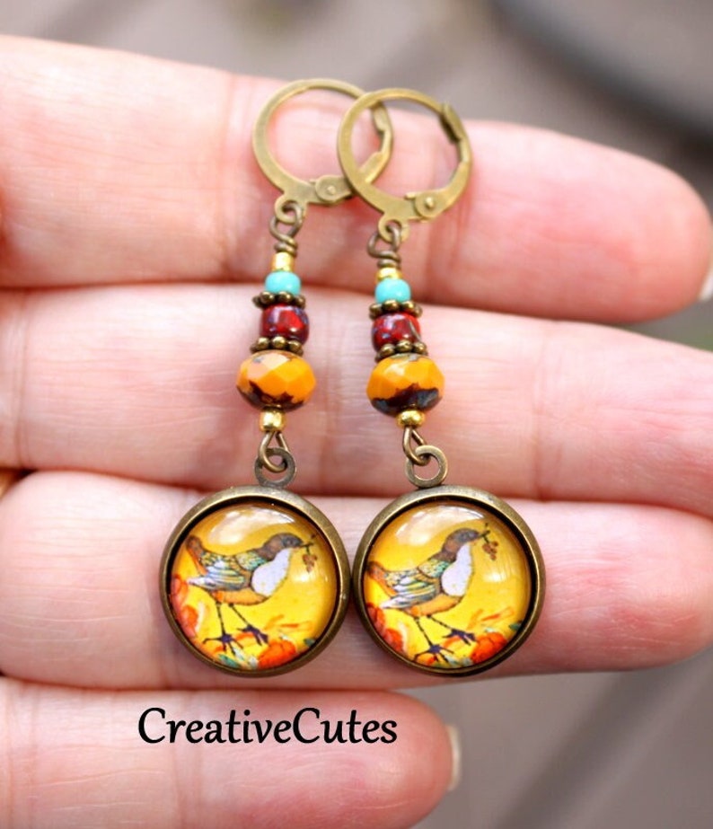 May include: A pair of dangle earrings with a bird and pumpkin design. The earrings feature a round, yellow glass cabochon with a bird and pumpkin image. The cabochon is attached to a bronze-colored chain with three beads: turquoise, red, and orange. The earrings are finished with bronze-colored hooks.