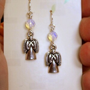 May include: A pair of silver angel earrings with iridescent white beads. The earrings are on a white card with the text "Creative Cutes Designs" and "www.CreativeCutes.Etsy.com".