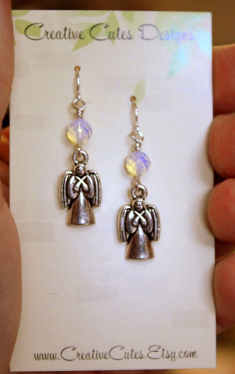 May include: A pair of silver angel earrings with iridescent white beads. The earrings are on a white card with the text "Creative Cutes Designs" and "www.CreativeCutes.Etsy.com".