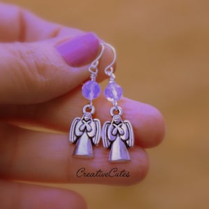 May include: Silver angel dangle earrings with a light purple bead on each earring. The earrings are held in a hand with purple nail polish.