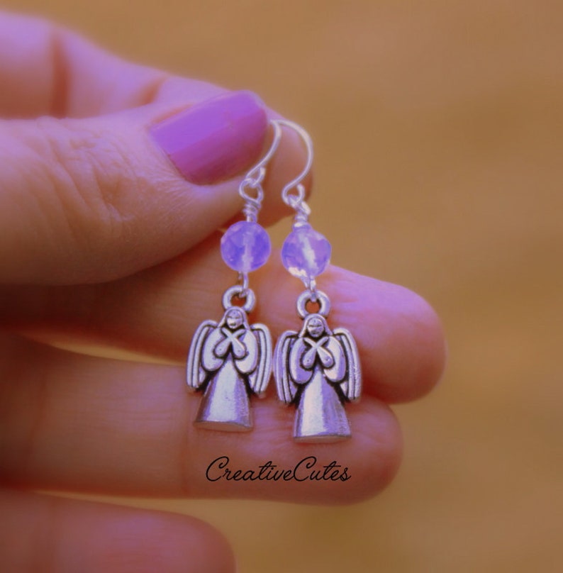 May include: Silver angel dangle earrings with a light purple bead on each earring. The earrings are held in a hand with purple nail polish.