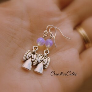 May include: Silver angel earrings with lavender iridescent glass beads. The earrings are on a person's hand.