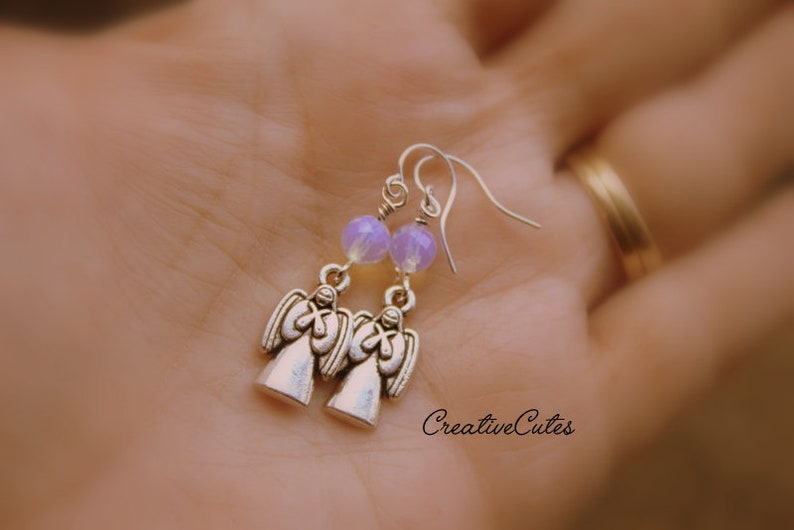 May include: Silver angel earrings with lavender iridescent glass beads. The earrings are on a person's hand.