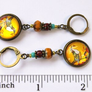 May include: A pair of gold tone earrings with a yellow and orange glass cabochon featuring a bird on a branch. The earrings are accented with turquoise and red beads and measure approximately 2 inches in length.