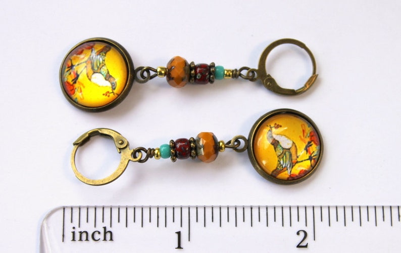 May include: A pair of gold tone earrings with a yellow and orange glass cabochon featuring a bird on a branch. The earrings are accented with turquoise and red beads and measure approximately 2 inches in length.