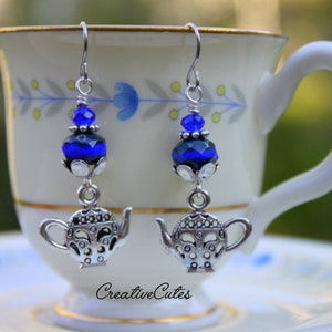 Peut inclure: Boucles d'oreilles pendantes en forme de théière argentée avec des perles de verre bleues et noires. Les boucles d'oreilles sont suspendues à une tasse à thé blanche.