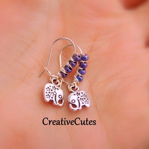 May include: A pair of silver elephant charm earrings with purple beads. The earrings are on a hand.