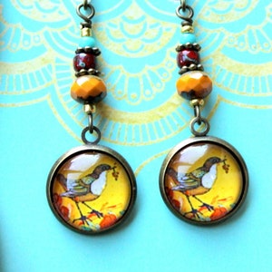 May include: A pair of earrings with a bird and floral design on a turquoise background. The earrings are made with glass cabochons, beads, and bronze findings. The earrings are handmade and feature a unique design.