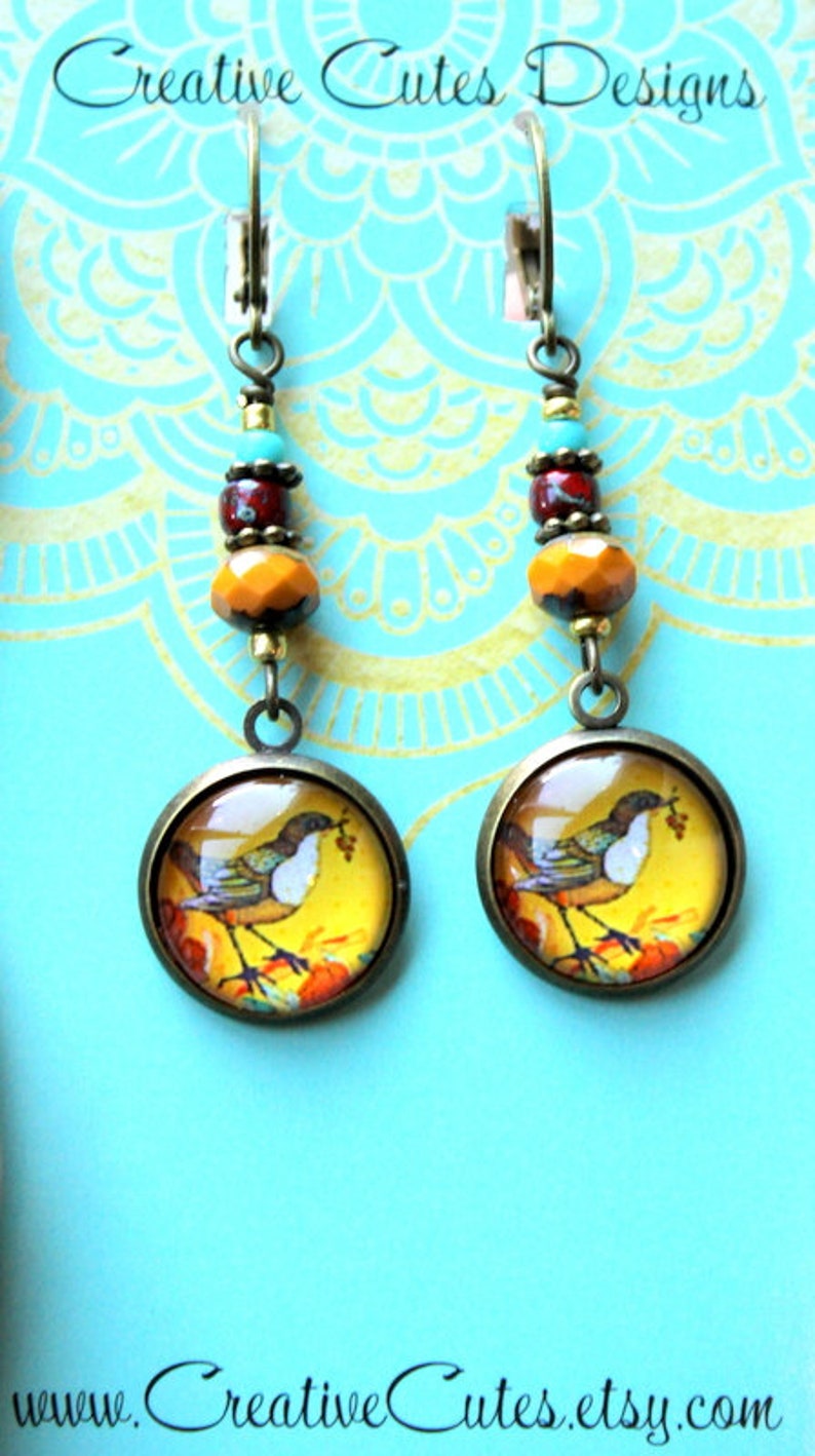 May include: A pair of earrings with a bird and floral design on a turquoise background. The earrings are made with glass cabochons, beads, and bronze findings. The earrings are handmade and feature a unique design.