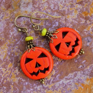 Könnte beinhalten: Ein Paar orangefarbene Ohrringe in Kürbisform mit einem geschnitzten Jack-o'-Lantern-Gesicht. Die Ohrringe sind aus bronzefarbenem Metall gefertigt und haben gelbe Perlen.