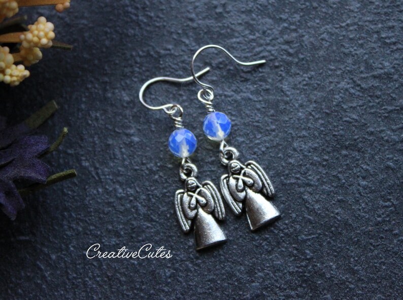 May include: Silver angel earrings with blue opal beads. The angels are facing forward with their wings outstretched. The earrings are hanging from silver hooks.
