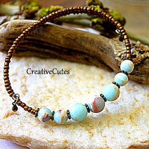 May include: A wrap bracelet featuring a long strand of small metallic bronze seed beads and a section of larger, round, light blue and tan marbled stone beads. A small bronze charm hangs from one end, and the text CreativeCutes is visible.