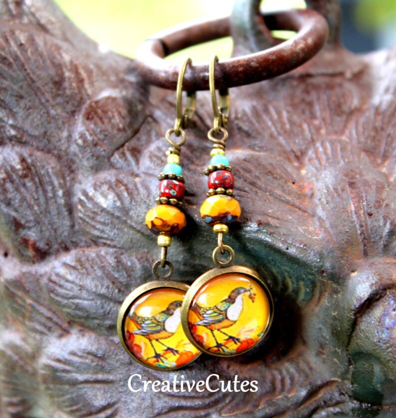 May include: A pair of dangle earrings with a bird design on the bottom. The earrings are made with gold-toned metal and feature a mix of yellow, red, and turquoise beads.