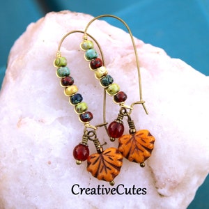 May include: A pair of gold hoop earrings with a colorful beaded design and a dangling leaf charm. The beads are in shades of green, blue, red, and yellow. The leaves are orange and brown.