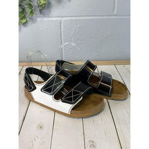 May include: A pair of sandals featuring a two-tone design with a white front strap and black heel strap, both accented by white contrast stitching. The sandals have adjustable hook-and-loop closures and a brown cork-textured footbed with a rubber sole.