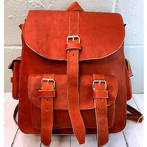 May include: A reddish-brown leather backpack with a flap closure and two front pockets. The backpack features adjustable straps and silver-colored buckles. The bag has a top handle and side pockets. The leather has a textured appearance.