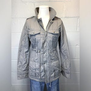 May include: A light gray jacket with a stand-up collar, four front pockets, and a drawstring waist. The jacket has a quilted pattern and is displayed on a mannequin. The jacket has a zipper and button closure.