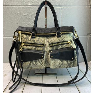 May include: A black and snakeskin-patterned handbag with gold-tone hardware. The bag features a braided top handle, multiple zippered compartments, and a detachable shoulder strap. The design includes black leather accents and gold zippers.