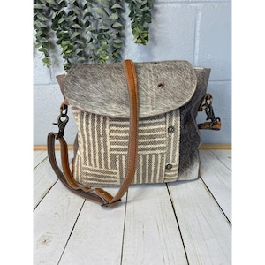 May include: A shoulder bag featuring a gray and white cowhide flap and a woven fabric body with a geometric striped pattern. The bag includes a brown leather strap, metal hardware, and two circular metal snap closures on the front panel.