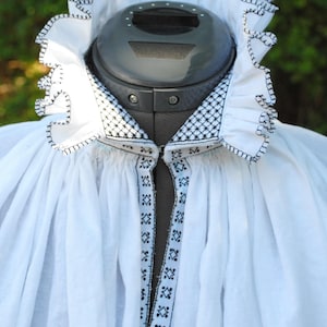 Puede incluir: Blusa blanca con cuello de volantes y ribete bordado en negro. El cuello tiene un patrón geométrico.