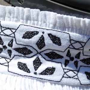 May include: Close-up of a white garment collar with a black embroidered floral design. The collar has a ruffled edge and a decorative band with geometric and floral patterns. The embroidery is detailed and adds a touch of elegance.