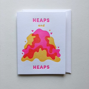 May include: A white card with the text "HEAPS and HEAPS" in pink and gold lettering. A pink and gold abstract design with hearts is in the center of the card.