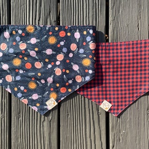 May include: Two dog bandanas, one with a black background and a pattern of planets and stars, and the other with a red and black checkered pattern.