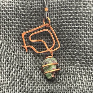 May include: A copper wire pendant with a green gemstone wrapped in copper wire. The pendant is hanging from a black cord.