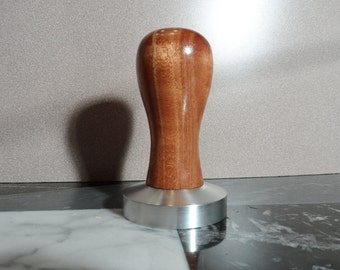 Könnte beinhalten: Ein Holztamper mit glatter, brauner Oberfläche und einer Basis aus Edelstahl. Der Tamper wird verwendet, um Kaffeepulver in einem Siebträger zu pressen.