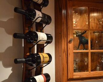 Puede incluir: Un estante de vino de madera con seis botellas de vino. El estante está montado en una pared y tiene una forma curva. Las botellas son de diferentes colores y tienen etiquetas.