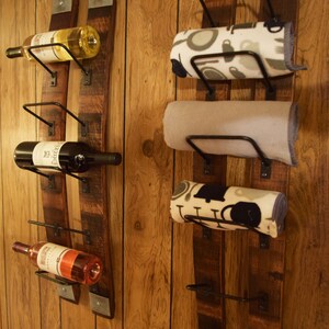 K&ouml;nnte beinhalten: Rustikales Weinregal und Handtuchhalter-Set. Das Weinregal enth&auml;lt drei Weinflaschen in verschiedenen Farben. Der Handtuchhalter hat drei aufgerollte Handt&uuml;cher mit verschiedenen Mustern. Beide sind an Holzbrettern an einer Holzvert&auml;felung befestigt.