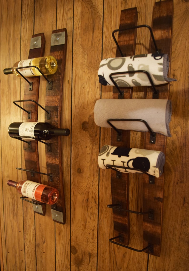 K&ouml;nnte beinhalten: Rustikales Weinregal und Handtuchhalter-Set. Das Weinregal enth&auml;lt drei Weinflaschen in verschiedenen Farben. Der Handtuchhalter hat drei aufgerollte Handt&uuml;cher mit verschiedenen Mustern. Beide sind an Holzbrettern an einer Holzvert&auml;felung befestigt.