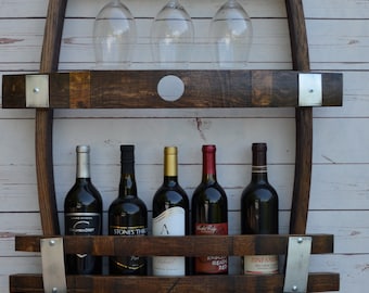 May include: A wooden wine rack with two shelves, featuring a circular cutout on the top shelf. The rack is mounted on a white wall and holds six bottles of wine and three wine glasses. The wine bottles have labels with various names, including "Columbia Crest", "Stones Throw", "Knocking Heads Red", and "Zinfandel".