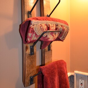 K&ouml;nnte beinhalten: Rustikaler Handtuchhalter aus Holz mit zwei Ebenen. Der Halter hat einen dunklen Metallrahmen und h&auml;lt zwei rote Handt&uuml;cher, eines gefaltet und das andere h&auml;ngend. Das gefaltete Handtuch hat eine dekorative Bord&uuml;re.