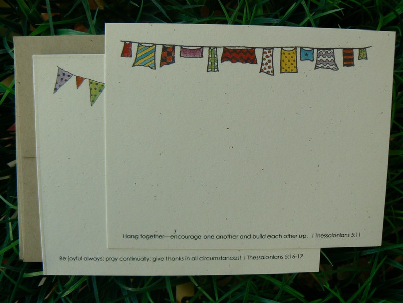 Op de afbeelding: Cr&egrave;mekleurige briefpapier set met een handgetekende illustratie van kleurrijke vlaggen en waslijnen. De set bevat notecards, enveloppen en een banner met hangende kledingstukken. De tekst luidt: "Hang together..." en "Be joyful always..."