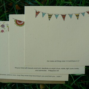 Op de afbeelding: Een stapel cr&egrave;mekleurige briefkaarten met decoratieve elementen en tekst. De bovenste kaart heeft een slinger van kleurrijke vlaggetjes. De tekst bevat bijbelverzen. De kaarten liggen op groen gras.