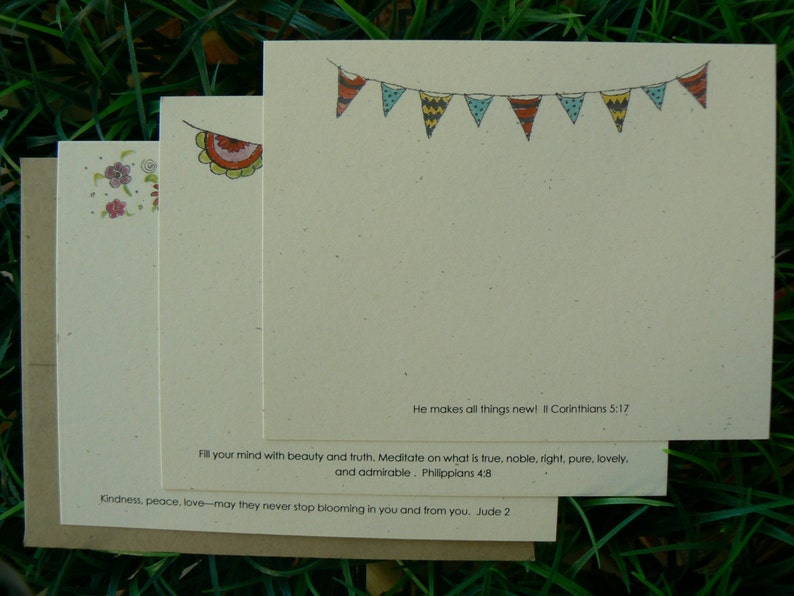 Op de afbeelding: Een stapel cr&egrave;mekleurige briefkaarten met decoratieve elementen en tekst. De bovenste kaart heeft een slinger van kleurrijke vlaggetjes. De tekst bevat bijbelverzen. De kaarten liggen op groen gras.