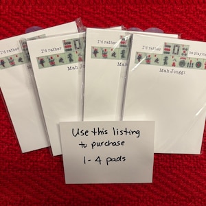 Puede incluir: Varios blocs de notas blancos, cada uno con una tira decorativa con fichas de Mahjong y la frase "I'd rather...be playing Mah Jongg!" La imagen incluye una nota que dice "Use this listing to purchase 1-4 pads."
