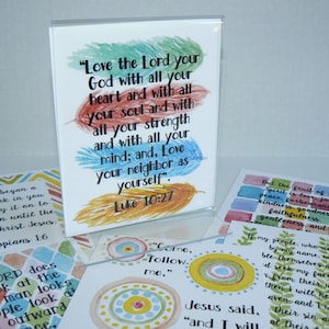 May include: A clear acrylic stand holds a white card featuring watercolor feathers in green, red, blue, and yellow with the black text: Love the Lord your God with all your heart and with all your soul and with all your strength and with all your mind; and, Love your neighbor as yourself. Luke 10:27. Several other cards with religious text and colorful watercolor designs are arranged around the base.