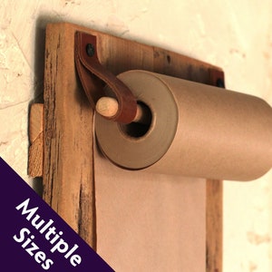 May include: A rustic wooden wall-mounted paper roll holder with a brown leather strap and a wooden dowel. The holder is made from reclaimed wood and has a natural, weathered finish.