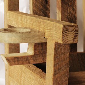 May include: Close-up of a wooden chair armrest with a built-in cup holder. The chair is constructed from rough-hewn, light-colored wood, showcasing the natural grain and texture. The cup holder is a circular indentation carved into the armrest.