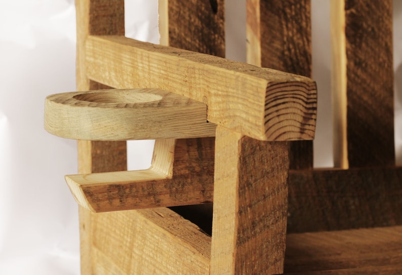 May include: Close-up of a wooden chair armrest with a built-in cup holder. The chair is constructed from rough-hewn, light-colored wood, showcasing the natural grain and texture. The cup holder is a circular indentation carved into the armrest.