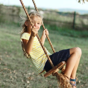 May include: A young person on a wooden swing with rope supports. The person wears a yellow and white striped shirt, navy shorts, and blue and orange shoes. The swing is in a grassy area with a blurred background.