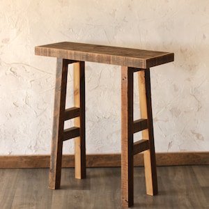 May include: A rustic, wooden console table with a rectangular top and four sturdy legs. The table is made of reclaimed wood, showcasing natural grain and knots. The legs are connected by horizontal supports, adding to the table's stability. The table measures approximately 48 inches long.