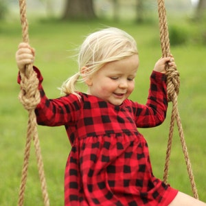 May include: A young child wearing a red and black plaid dress is smiling while sitting on a wooden swing. The swing is suspended by thick, knotted ropes. The background is a blurred green field and trees.